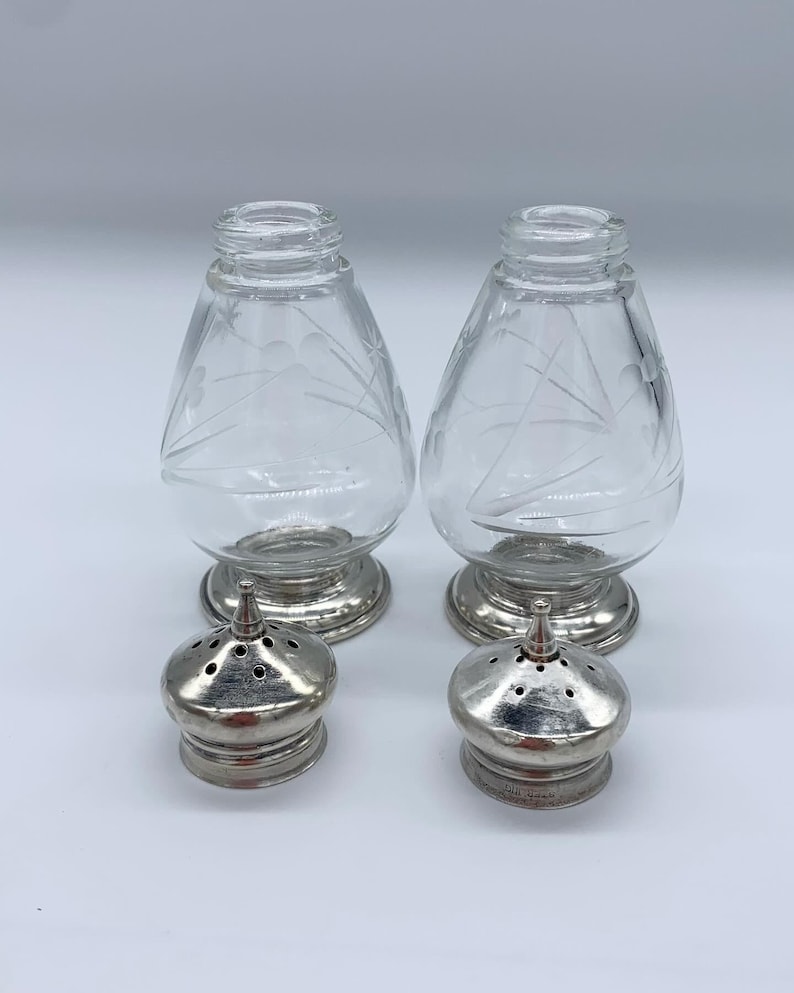 May include: A set of two clear glass salt and pepper shakers with etched designs. The shakers have silver tops with a small hole for dispensing salt and pepper.