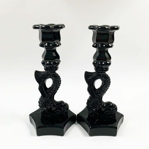 May include: A pair of black glass candlestick holders shaped like a mermaid with a tail and a fin. The holders have a hexagonal base.