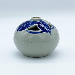 May include: A round, ceramic vase with a light grey base and blue leaf designs. The vase has three triangular openings and a small opening at the top. The blue leaf design is painted on the top of the vase.