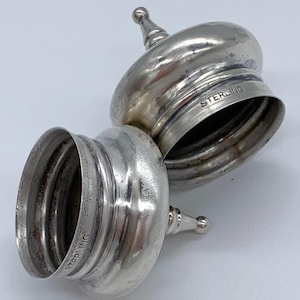 May include: Two silver-toned, round, lidded containers with a domed top and a small knob on top. The lids are marked "STERLING" on the bottom.