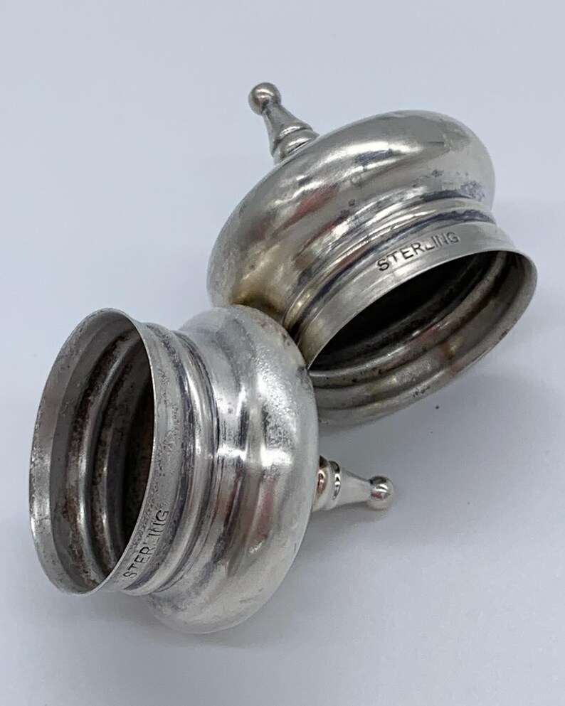 May include: Two silver-toned, round, lidded containers with a domed top and a small knob on top. The lids are marked "STERLING" on the bottom.