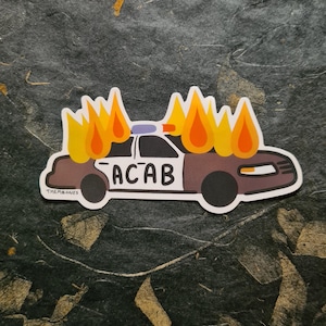 May include: A black and white sticker of a police car on fire with the text "ACAB" on the side. The sticker is on a dark gray textured background.