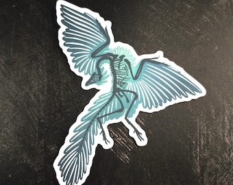 May include: A die-cut sticker featuring a stylized illustration of a bird skeleton in flight. The skeletal structure is rendered in dark gray lines against a soft, light blue glowing aura, with large, detailed wings spread wide.