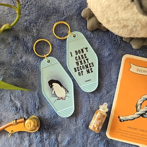 May include: Two light blue keychains with gold rings. One keychain features a penguin illustration, the other reads "I DON'T CARE WHAT BECOMES OF ME." A small bottle with gold glitter and a card are also visible.