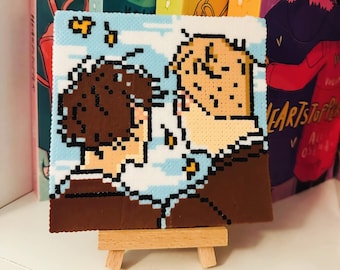 Könnte beinhalten: Ein Pixel-Kunstwerk auf einem kleinen Holzstaffelei, das zwei Personen zeigt, die sich ansehen. Der Hintergrund ist ein blauer Himmel mit weißen Wolken. Die Personen sind braun und gelb. Das Werk befindet sich in einem Regal mit anderen Büchern.