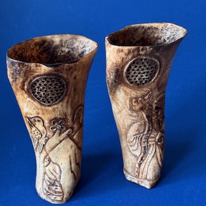 May include: Two carved horn vessels with a light tan, textured surface. Each vessel features a circular, honeycomb-patterned inlay near the top rim. The left vessel displays etched bird and floral motifs, while the right vessel shows a figure in robes.