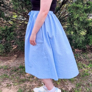 May include: A light blue midi skirt with a gathered waist and a black top. The skirt is paired with white sneakers with blue accents. The outfit is worn outdoors with a green background.