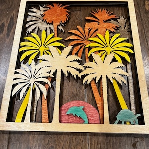 May include: A wooden wall art piece featuring a layered design of palm trees in shades of yellow, orange, and brown. A dolphin and a turtle are cut out of the wood and placed in front of the trees. The piece is framed in a natural wood frame.