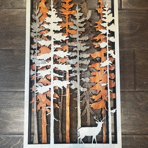 Könnte beinhalten: Ein mehrschichtiges Wandkunstwerk aus Holz, das eine Waldszene mit Bäumen in Weiß-, Braun- und Orangetönen darstellt. Eine weiße Silhouette eines Hirsches befindet sich im Vordergrund. Das Kunstwerk ist in einem weißen und schwarzen Rahmen eingefasst.