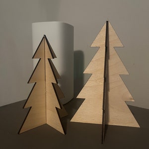 Puede incluir: Dos adornos de &aacute;rbol de Navidad de madera, uno m&aacute;s grande que el otro, ambos de color marr&oacute;n claro y dise&ntilde;o sencillo.
