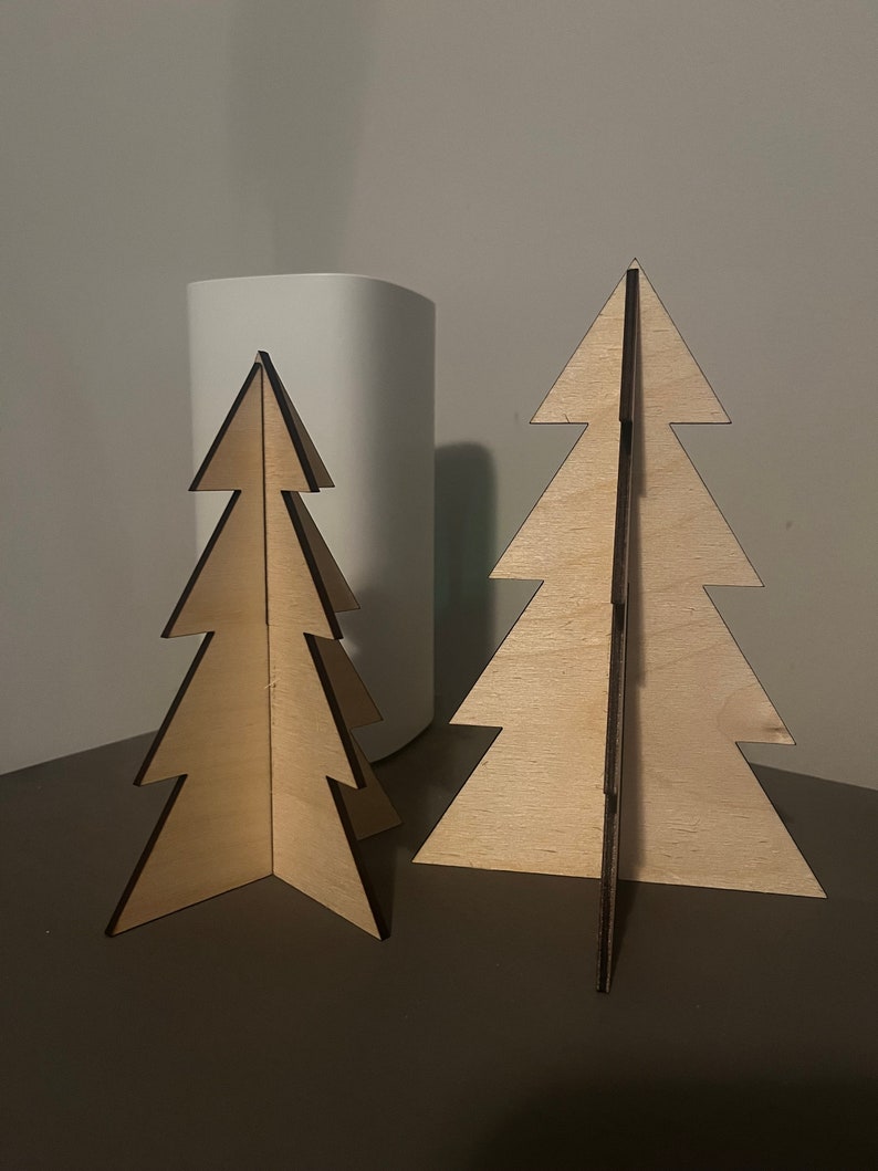Puede incluir: Dos adornos de &aacute;rbol de Navidad de madera, uno m&aacute;s grande que el otro, ambos de color marr&oacute;n claro y dise&ntilde;o sencillo.