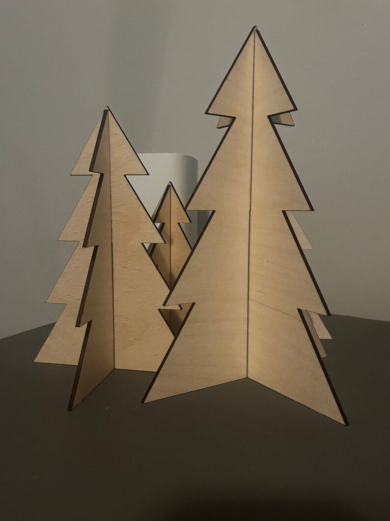 Puede incluir: Tres &aacute;rboles de Navidad de madera de diferentes tama&ntilde;os, de pie sobre una superficie gris oscuro. Los &aacute;rboles est&aacute;n hechos de madera contrachapada y tienen un acabado de madera natural.