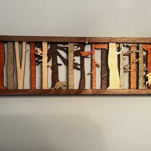 May include: A wooden wall art piece depicting a forest scene with trees, a deer, and a bear. The piece is made of different shades of wood, creating a layered effect.