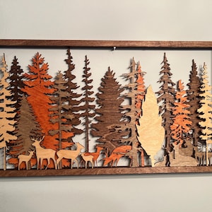 Puede incluir: Una pieza de arte de pared de madera que representa una escena de bosque con múltiples siluetas de árboles y ciervos. Los árboles están cortados en madera y pintados en tonos de marrón, naranja y beige. Los ciervos también están cortados en madera y pintados en tonos de marrón.