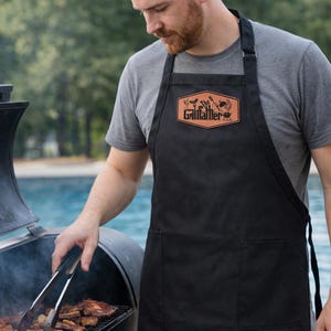 May include: Black apron with two front pockets and the text "Grillfather" on a brown patch. The apron is worn by a person grilling meat on a barbecue. The person is using tongs to handle the food.