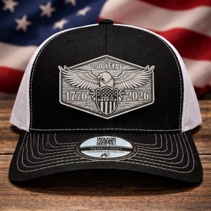 May include: Black and white trucker hat with a patch commemorating 250 years of America. The patch features an eagle, the years 1776 and 2026, and the words "250 Years" and "Made in America". The hat has a curved brim and a white mesh back.
