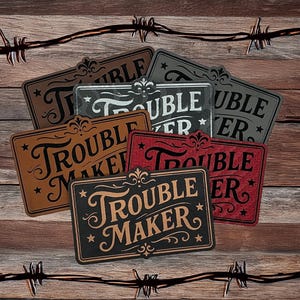 May include: A collection of rectangular signs with the words "Trouble Maker" in a decorative font. The signs are in various colors, including brown, black, and red, with a rustic design. The background is a wooden surface with barbed wire.