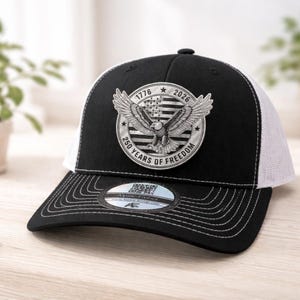 May include: Black and white trucker hat with a patch featuring an eagle, American flag, and the text "1776 2026 250 Years of Freedom." The hat has a curved brim and a mesh back.