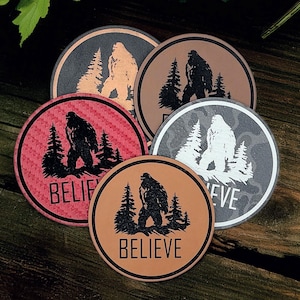 May include: A collection of round coasters featuring a Bigfoot silhouette with pine trees. The coasters are in various colors, including red, brown, and gray, with the word "BELIEVE" or "EVE" printed on them. The coasters are on a wooden surface.