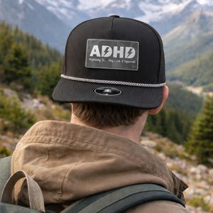 May include: Black trucker hat with a white and silver rope detail. The hat features a rectangular patch with the letters "ADHD" and the phrase "Highway To... Hey Look A Squirrel" in white text. The hat is worn outdoors.