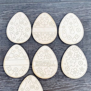 Può includere: Sette forme di uova di Pasqua in legno non finite con vari disegni. Alcune uova hanno disegni di coniglietti e fiori, mentre altre hanno uno spazio vuoto per la personalizzazione. Un uovo ha il nome "KENDALL" scritto su di esso.