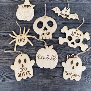 May include: Nine wooden Halloween ornaments with names on them. The ornaments are shaped like a pumpkin, bat, spider, skull, skull and crossbones, apple, and two ghosts.