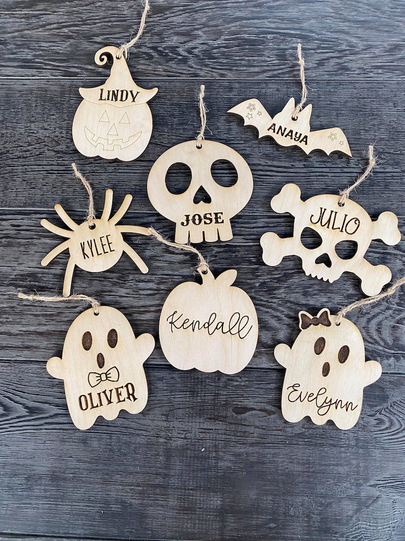 May include: Nine wooden Halloween ornaments with names on them. The ornaments are shaped like a pumpkin, bat, spider, skull, skull and crossbones, apple, and two ghosts.