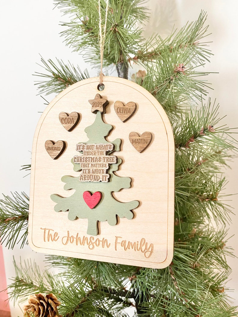 Peut inclure: Une d&eacute;coration de No&euml;l en bois en forme d'arbre avec le texte "Ce n'est pas ce qui se trouve sous le sapin de No&euml;l qui compte, mais qui est autour." L'ornement comporte &eacute;galement une &eacute;toile, des c&oelig;urs avec des noms et l'ann&eacute;e 2023. Le bas de l'ornement porte l'inscription "The Johnson Family".