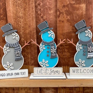 Puede incluir: Tres figuras de mu&ntilde;eco de nieve de madera con cuerpos de diferentes colores. El mu&ntilde;eco de nieve de la izquierda es gris con un sombrero y una bufanda negros. El mu&ntilde;eco de nieve del medio es azul con un sombrero y una bufanda negros. El mu&ntilde;eco de nieve de la derecha es azul verdoso con un sombrero y una bufanda negros. Cada mu&ntilde;eco de nieve tiene un letrero delante. El letrero de la izquierda dice "A&ntilde;ade tu propio texto". El letrero del medio dice "Que nieve". El letrero de la derecha dice "Bienvenido".