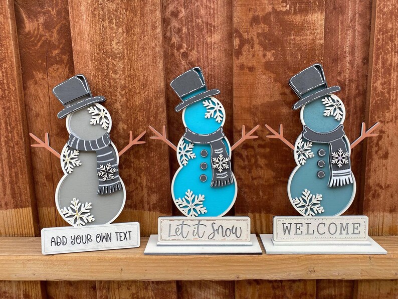 Puede incluir: Tres figuras de mu&ntilde;eco de nieve de madera con cuerpos de diferentes colores. El mu&ntilde;eco de nieve de la izquierda es gris con un sombrero y una bufanda negros. El mu&ntilde;eco de nieve del medio es azul con un sombrero y una bufanda negros. El mu&ntilde;eco de nieve de la derecha es azul verdoso con un sombrero y una bufanda negros. Cada mu&ntilde;eco de nieve tiene un letrero delante. El letrero de la izquierda dice "A&ntilde;ade tu propio texto". El letrero del medio dice "Que nieve". El letrero de la derecha dice "Bienvenido".