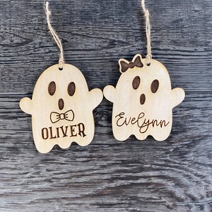 May include: Two wooden ghost ornaments with names "Oliver" and "Evelynn" written on them. The ghost ornaments are hanging from twine.
