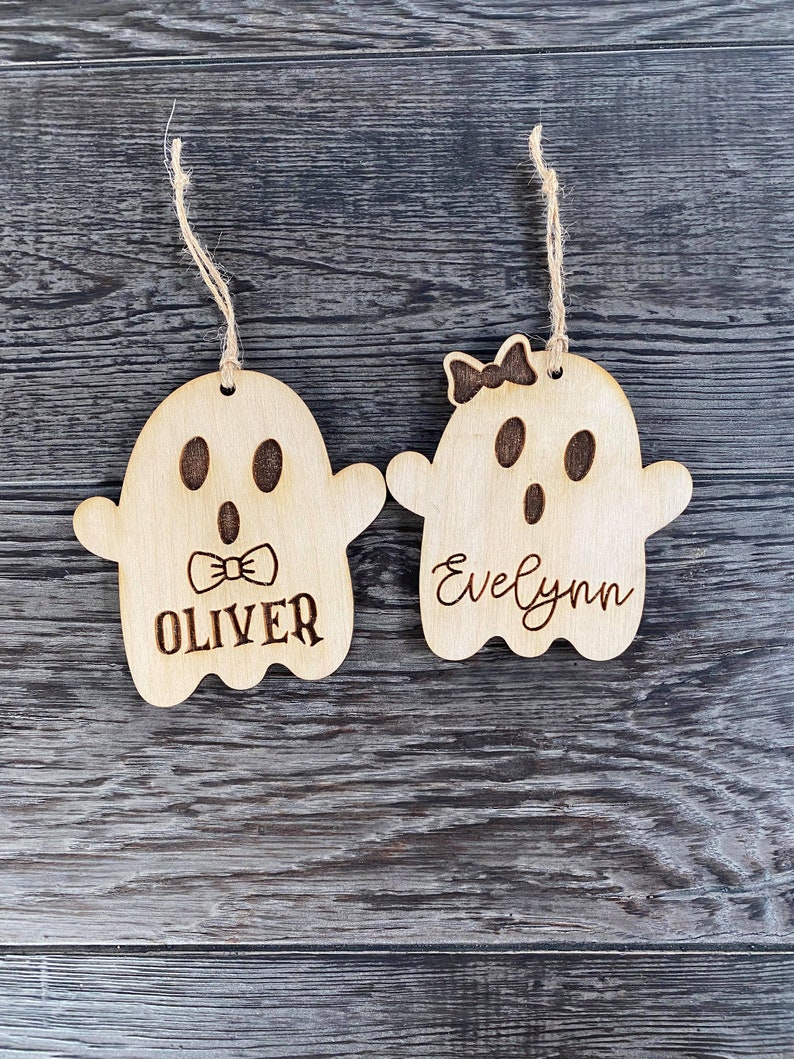 May include: Two wooden ghost ornaments with names "Oliver" and "Evelynn" written on them. The ghost ornaments are hanging from twine.