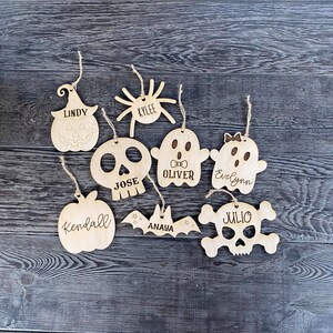May include: Eight wooden Halloween ornaments with different designs, including a pumpkin, spider, ghost, skull, bat, and a skull and crossbones. Each ornament has a name written on it, including Lindy, Kylee, Oliver, Evelyn, Jose, Kendall, Anaya, and Julio.