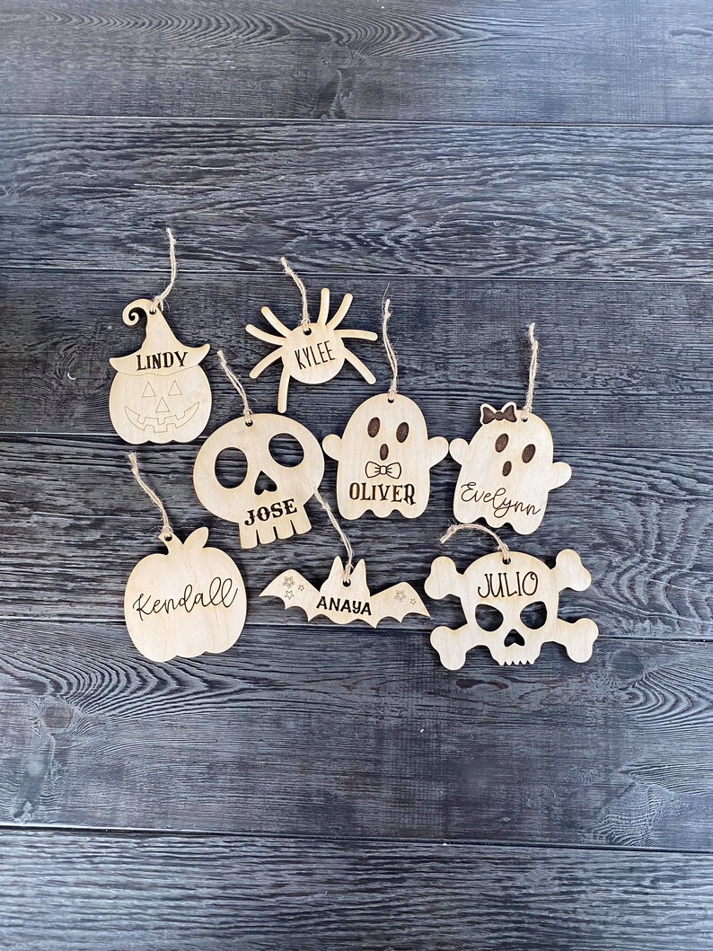 May include: Eight wooden Halloween ornaments with different designs, including a pumpkin, spider, ghost, skull, bat, and a skull and crossbones. Each ornament has a name written on it, including Lindy, Kylee, Oliver, Evelyn, Jose, Kendall, Anaya, and Julio.