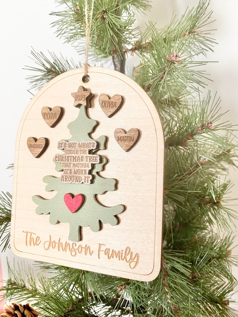 Peut inclure: Une d&eacute;coration de No&euml;l en bois en forme d'arbre avec le texte "Ce n'est pas ce qui se trouve sous le sapin de No&euml;l qui compte, mais qui est autour." et les noms "Antonio", "Cora", "Olivia", "Martin" et "The Johnson Family".