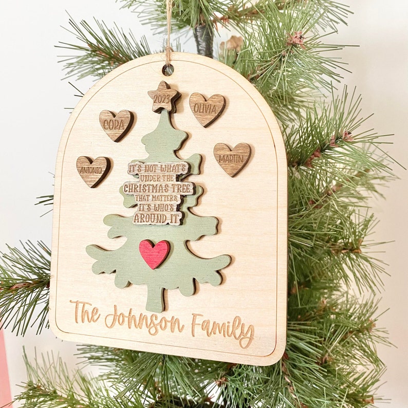 Peut inclure: Une d&eacute;coration de No&euml;l en bois en forme de sapin de No&euml;l avec le texte "Ce n'est pas ce qui se trouve sous le sapin de No&euml;l qui compte, c'est qui est autour." La d&eacute;coration porte &eacute;galement le texte "The Johnson Family" et cinq c&oelig;urs avec des noms dessus : Antonio, Cora, Olivia, Martin et 2023.