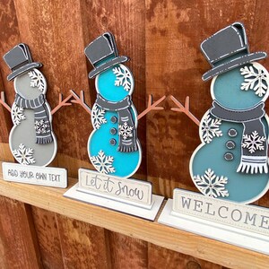 Puede incluir: Tres decoraciones de mu&ntilde;eco de nieve de madera con sombreros negros y copos de nieve blancos. Los mu&ntilde;ecos de nieve est&aacute;n pintados en tonos de azul y gris. Los letreros debajo de los mu&ntilde;ecos de nieve dicen "A&ntilde;ade tu propio texto", "Deja que nieve" y "Bienvenido".