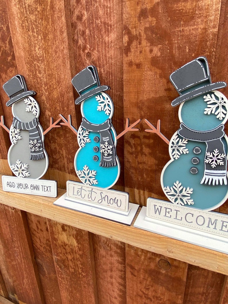 Puede incluir: Tres decoraciones de mu&ntilde;eco de nieve de madera con sombreros negros y copos de nieve blancos. Los mu&ntilde;ecos de nieve est&aacute;n pintados en tonos de azul y gris. Los letreros debajo de los mu&ntilde;ecos de nieve dicen "A&ntilde;ade tu propio texto", "Deja que nieve" y "Bienvenido".