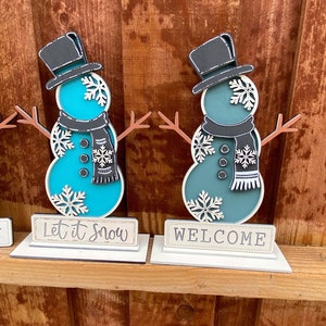 Puede incluir: Dos figuras de mu&ntilde;eco de nieve de madera con sombreros y bufandas negros, de pie sobre bases blancas con letreros que dicen "Let it Snow" y "Welcome".