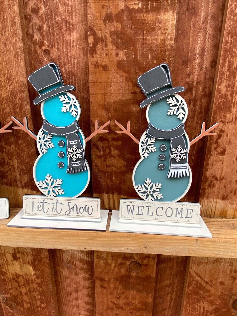 Puede incluir: Dos figuras de mu&ntilde;eco de nieve de madera con sombreros y bufandas negros, de pie sobre bases blancas con letreros que dicen "Let it Snow" y "Welcome".