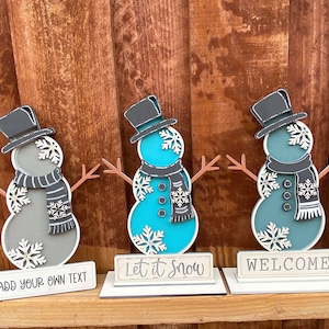 Puede incluir: Tres figuras de mu&ntilde;eco de nieve de madera con sombreros y bufandas negros. El mu&ntilde;eco de nieve de la izquierda es gris, el del medio es turquesa y el de la derecha es azul verdoso. Cada mu&ntilde;eco de nieve tiene un letrero delante. El letrero de la izquierda dice "A&ntilde;ade tu propio texto", el del medio dice "Deja que nieve" y el de la derecha dice "Bienvenido".