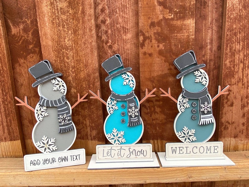 Puede incluir: Tres figuras de mu&ntilde;eco de nieve de madera con sombreros y bufandas negros. El mu&ntilde;eco de nieve de la izquierda es gris, el del medio es turquesa y el de la derecha es azul verdoso. Cada mu&ntilde;eco de nieve tiene un letrero delante. El letrero de la izquierda dice "A&ntilde;ade tu propio texto", el del medio dice "Deja que nieve" y el de la derecha dice "Bienvenido".