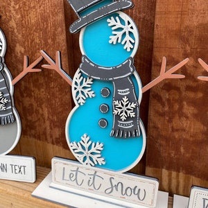 Puede incluir: Una figura de mu&ntilde;eco de nieve azul y blanco con un sombrero de copa negro y una bufanda. El mu&ntilde;eco de nieve est&aacute; de pie sobre una base blanca con el texto "Let it Snow" escrito en un cartel.