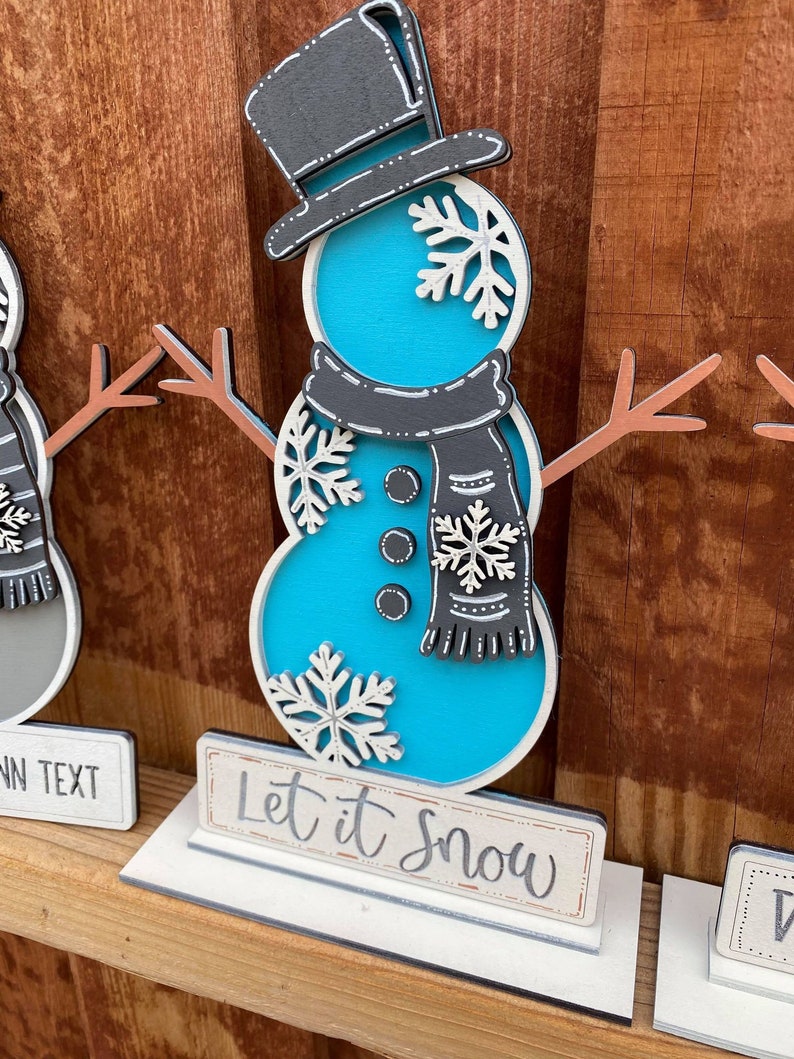 Puede incluir: Una figura de mu&ntilde;eco de nieve azul y blanco con un sombrero de copa negro y una bufanda. El mu&ntilde;eco de nieve est&aacute; de pie sobre una base blanca con el texto "Let it Snow" escrito en un cartel.