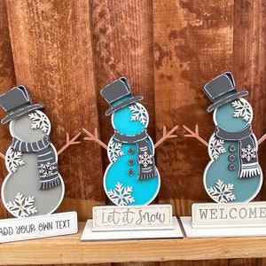 Puede incluir: Tres figuras de mu&ntilde;eco de nieve de madera con sombreros y bufandas negros. El mu&ntilde;eco de nieve de la izquierda es gris, el del medio es turquesa y el de la derecha es azul verdoso. Cada mu&ntilde;eco de nieve tiene un letrero delante. El letrero de la izquierda dice "A&ntilde;ade tu propio texto", el del medio dice "Deja que nieve" y el de la derecha dice "Bienvenido".