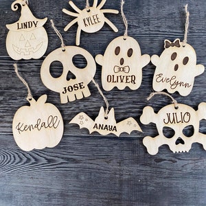 May include: Eight wooden Halloween ornaments with different designs and names. The ornaments include a pumpkin, a spider, a ghost, a skull, a bat, and a skull with crossbones. The names on the ornaments are Lindy, Kylee, Oliver, Evelyn, Jose, Anaya, Kendall, and Julio.