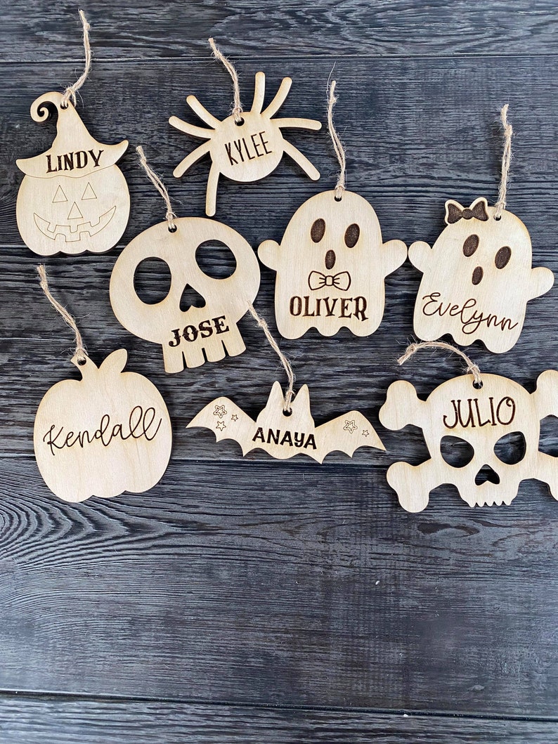 May include: Eight wooden Halloween ornaments with different designs and names. The ornaments include a pumpkin, a spider, a ghost, a skull, a bat, and a skull with crossbones. The names on the ornaments are Lindy, Kylee, Oliver, Evelyn, Jose, Anaya, Kendall, and Julio.