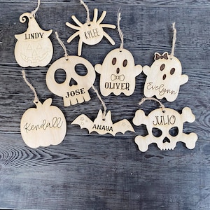 May include: Eight wooden Halloween ornaments with names on them. The ornaments are shaped like a pumpkin, spider, ghost, skull, bat, and a skull and crossbones.