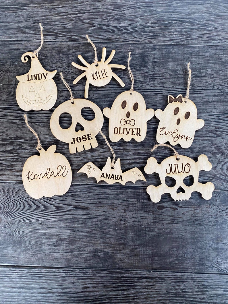 May include: Eight wooden Halloween ornaments with names on them. The ornaments are shaped like a pumpkin, spider, ghost, skull, bat, and a skull and crossbones.