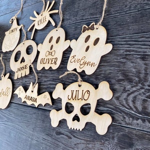 May include: A set of eight wooden Halloween ornaments with names written on them. The ornaments are shaped like a pumpkin, spider, ghost, skull, bat, and apple.